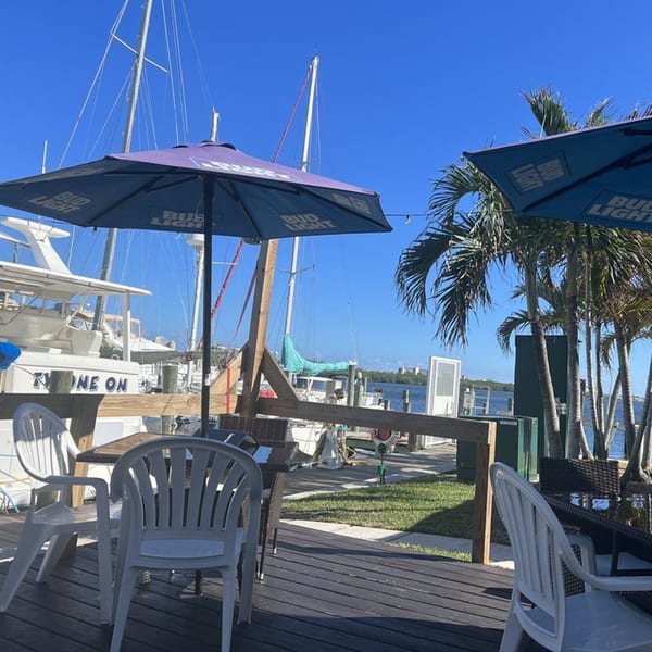 Outdoor dining area at Sauder's Landing with tables, umbrellas, palm trees, and boats docked by the water.
