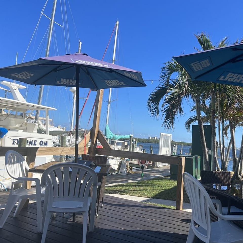 Outdoor dining area at Sauder's Landing with tables, umbrellas, palm trees, and boats docked by the water.