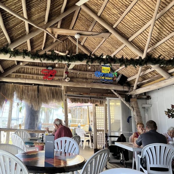 Diners seated at tables under a thatched roof with neon beer signs at Sauder's Landing Restaurant.