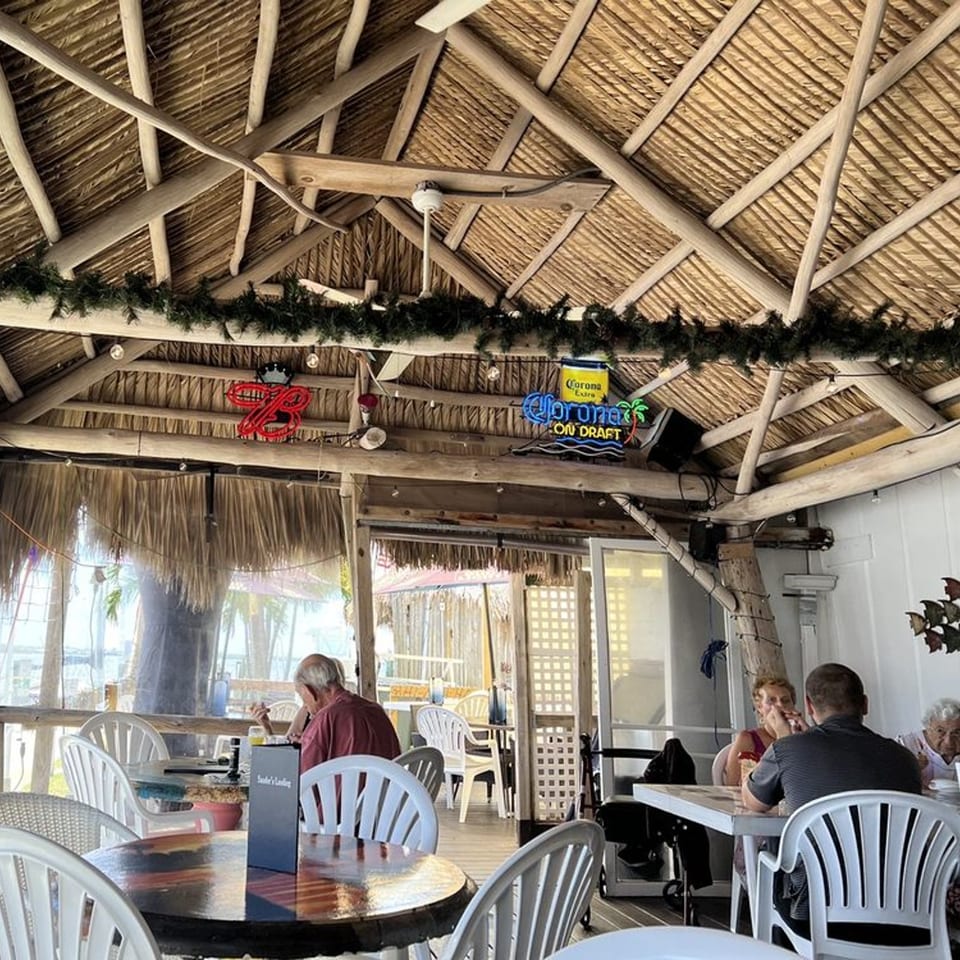 Diners seated at tables under a thatched roof with neon beer signs at Sauder's Landing Restaurant.