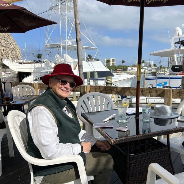 A smiling person in a red hat and sunglasses sits at an outdoor table overlooking a marina.